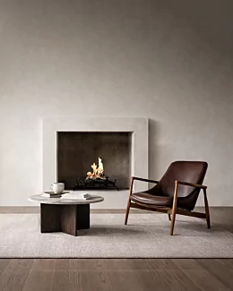 Modern minimalist living room featuring a built-in fireplace with a clean-burning flame set in a recessed concrete surround. In front, a round stone coffee table with books and a white mug sits on a textured rug beside a mid-century style brown leather lounge chair with wooden arms, creating a warm, contemporary interior setting.